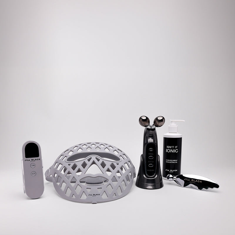 A skincare set on a light gray background, including a gray mesh head, a black facial cleansing device, a black bottle labeled SKIN ACID KINSE, a black spatula, and a gray remote control.