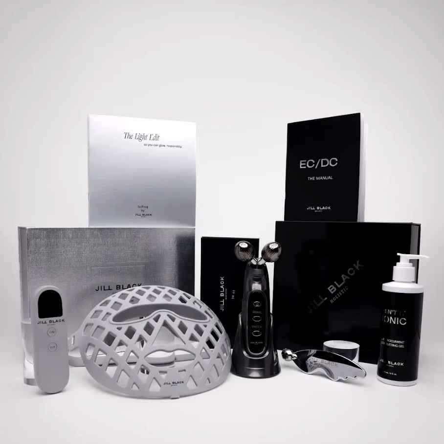 A JILL BLACK skincare set arranged on a light gray studio background, including a silver treatment helmet, black device, silver and black boxes, a white pump bottle labeled JILL BLACK CLINIC, and other included accessories.