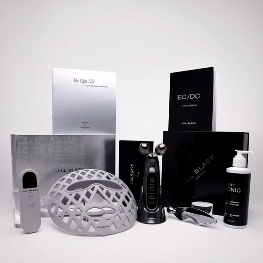 A JILL BLACK skincare set arranged on a light gray studio background, including a silver treatment helmet, black device, silver and black boxes, a white pump bottle labeled JILL BLACK CLINIC, and other included accessories.